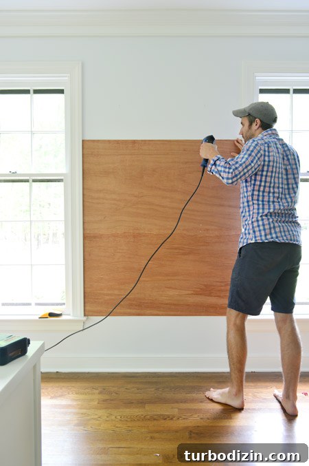 Create a Cork Wall Gallery for Kids’ Artwork 6 using power drill to screw plywood planks on wall between two windows to become base for gluing cork board tiles to create cork board wall