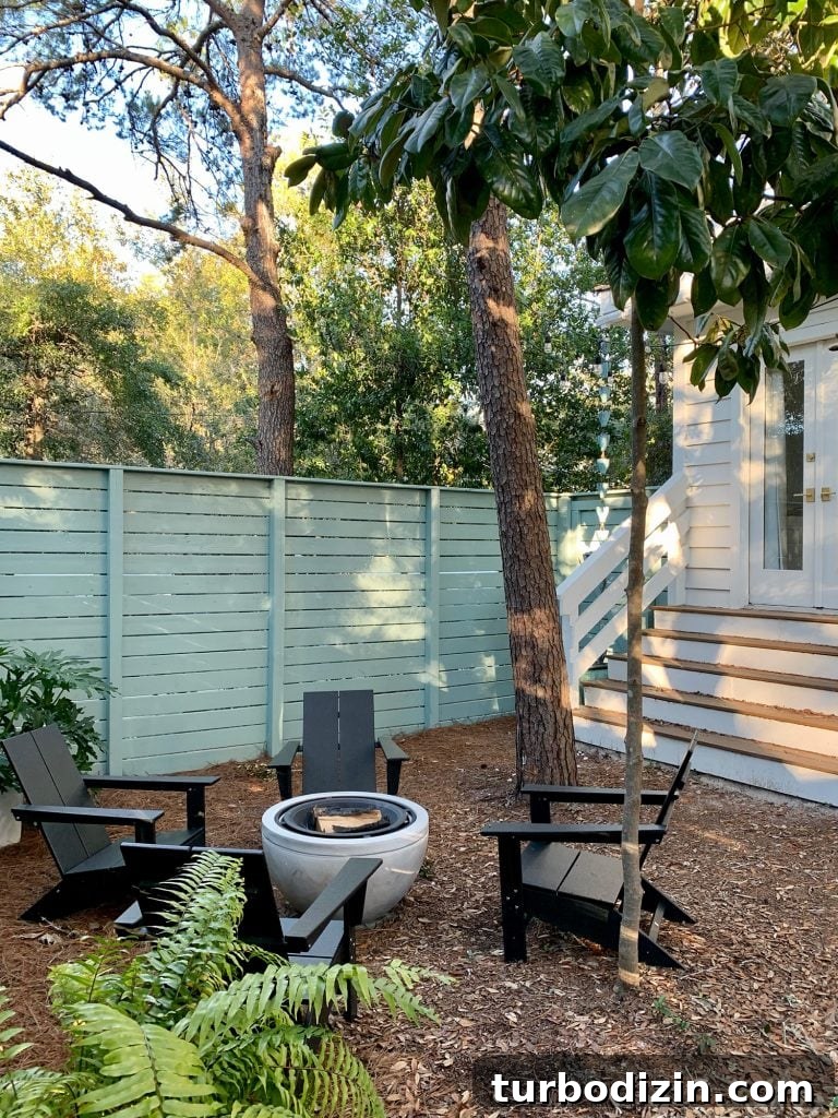 Trees around fenced side yard with black Adirondack chairs around fire pit