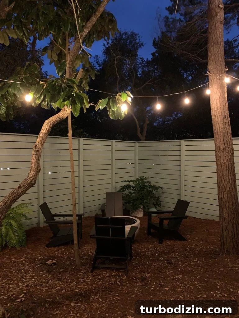 Nighttime view of fenced side yard with black Adirondack chairs around fire pit